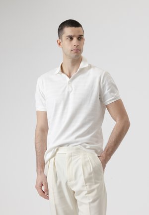 Hombre con camiseta polo blanca texturizada y pantalones crema plisados, de pie con una mano en el bolsillo, mirando ligeramente hacia la derecha.