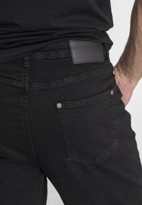 Jeans de mezclilla negra con una textura suave, que presentan una etiqueta de cuero en la cintura y herrajes dorados en los bolsillos traseros.