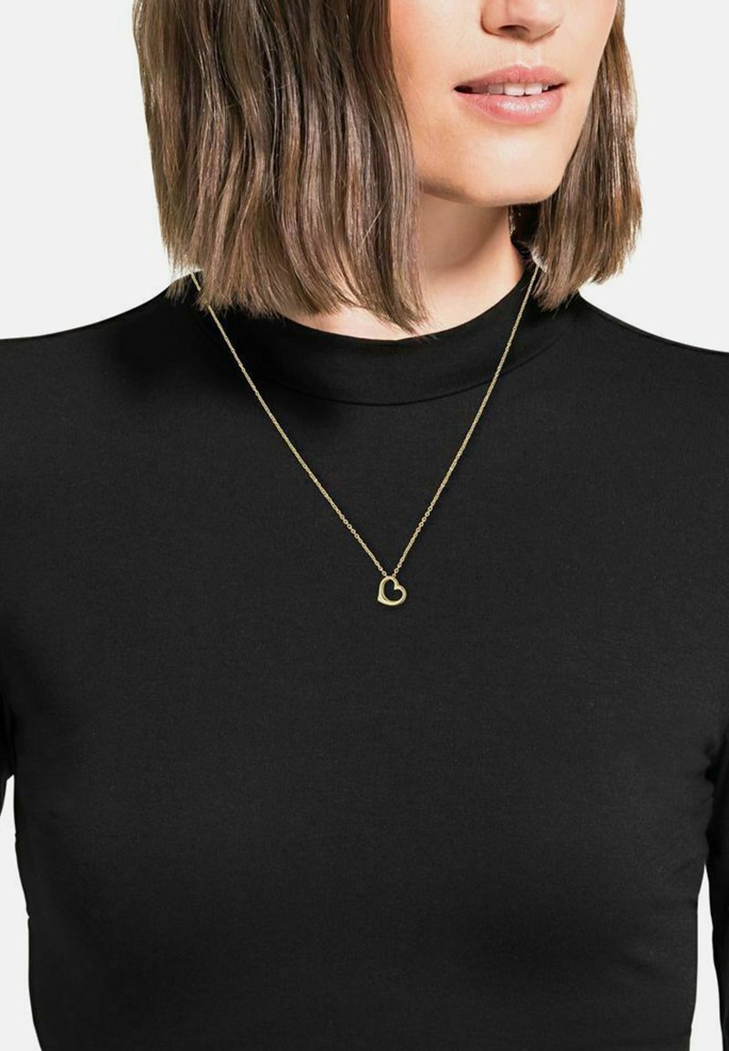 Collana con pendente a forma di cuore in oro su una delicata catena, indossata su un top nero a collo alto. Design semplice con una finitura liscia.