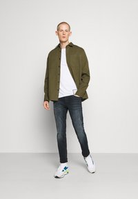 Camisa verde-oliva de botão sobre uma t-shirt branca, combinada com jeans escuros e ténis brancos com detalhes azuis e verdes.