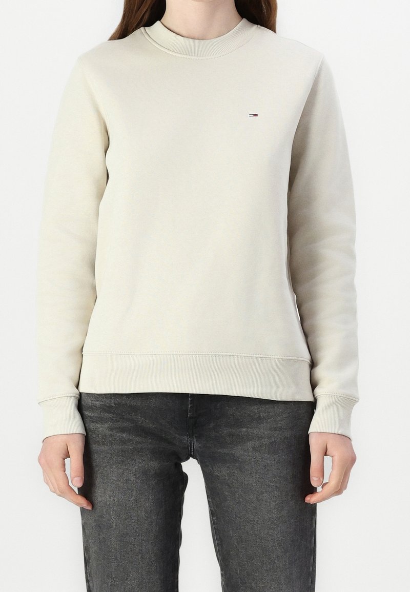 Felpa beige chiaro con collo tondo e polsi a coste. Presenta un piccolo logo multicolore sul petto. Abbinata a jeans grigi scuri.