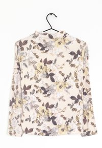 Top a maniche lunghe con colletto a imbuto e motivo floreale su sfondo beige, appeso a un gancio nero contro una parete bianca.