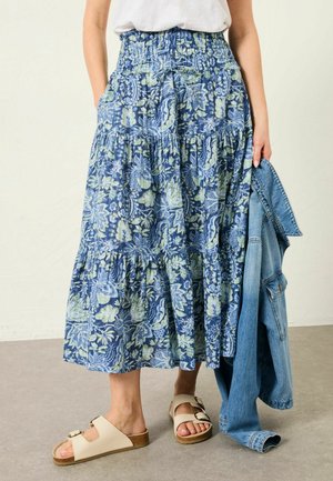 Blue floral-patterned maxi skirt with gathered waist, side pockets, and a tiered design. Paired with cream slide sandals and a denim jacket.