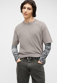 Νεαρό άτομο με μεσαίου μήκους σκούρα μαλλιά, που φοράει γκρι μπλουζάκι πάνω από μακρυμάνικη μπλούζα με ασπρόμαυρο ζεβρέ σχέδιο, σκούρο τζιν και μαύρη ζώνη.