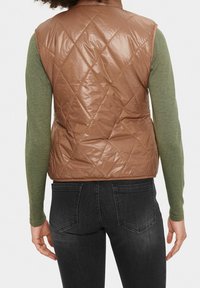Brązowa pikowana kamizelka o błyszczącej powierzchni, bez rękawów i z wysokim kołnierzem. Noszona na zielonej koszuli z długim rękawem i szarych dżinsach.
