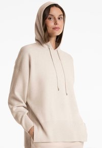 Jeune femme portant un sweat à capuche oversized beige à manches longues, les mains dans les poches, se tenant devant un fond blanc uni.