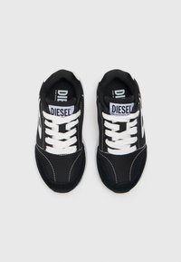 Fekete sportcipők fehér fűzőkkel és kiegészítőkkel, textúrált textil felsőrész és párnázott talp jellemzi, az orrán "DIESEL" felirattal.