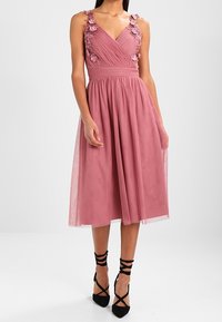 Vaaleanpunainen olkaimeton mekko, jossa on kukkakoristeet harteilla, tiukka yläosa ja laskeutuva hame, yhdistettynä mustiin remmi-korkokenkiin.