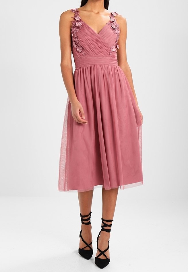 Vaaleanpunainen olkaimeton mekko, jossa on kukkakoristeet harteilla, tiukka yläosa ja laskeutuva hame, yhdistettynä mustiin remmi-korkokenkiin.