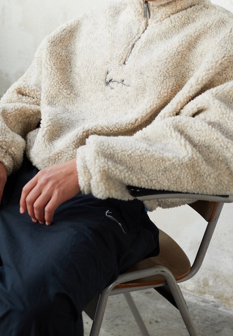 Cremefarbener Fleece-Pullover mit einem Viertel-Reißverschluss und Logostickerei, getragen über dunkler Hose, die Textur ist weich und flauschig.