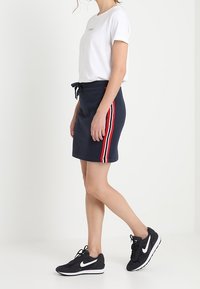 Donna in maglietta bianca, gonna blu navy con strisce laterali rosse e bianche, e sneakers nere Nike su sfondo bianco.