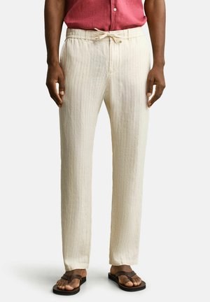 Mann trägt beige Nadelstreifenhose mit Kordelzug, rotes Hemd und braune Flip-Flops, steht vor einfarbigem Hintergrund.