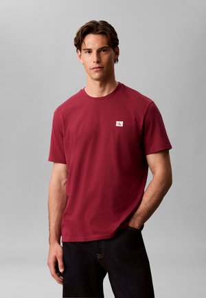 Jeune homme portant un t-shirt rouge uni avec un petit logo blanc sur la poitrine, debout avec une main dans la poche sur fond gris.