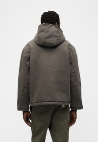 Άνδρας που φοράει σκούρο γκρι μπουφάν με κουκούλα και χακί παντελόνια, στέκεται με την πλάτη προς τα εμπρός σε απλό ανοιχτόχρωμο φόντο.