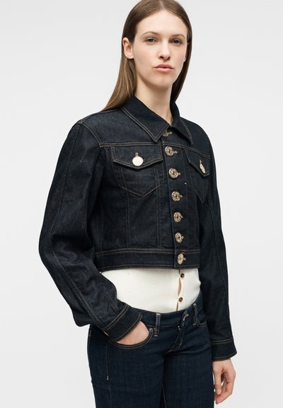 Γυναίκα με σκούρο τζιν μπουφάν με χρυσά κουμπιά πάνω από λευκό ριγωτό τοπ, συνδυασμένο με τζιν παντελόνι σε σκούρο τζιν χρώμα, το χέρι στην τσέπη.