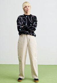 Sweater preto com padrão abstrato em branco e azul, combinado com calças beiges de cintura alta. Modelo em pé sobre uma superfície verde, usando sapatos pretos.