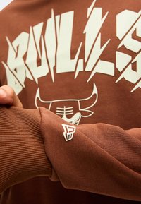 Καφέ φούτερ Chicago Bulls με έντονα λευκά γράμματα και λογότυπο ταύρου, μανίκι με ριμπ μανσέτα και μικρό λογότυπο ομάδας.