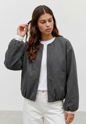 Jeune femme aux longs cheveux bruns ondulés portant une veste grise zippée avec des manches bouffantes et un pantalon blanc, sur un fond clair uni.