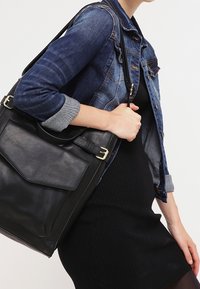 Svart läder tote-väska med en framficka, guldfärgade detaljer och en avtagbar axelrem, hängande p å axeln av en jeansjacka.