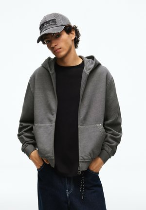 Junger Mann trägt einen grauen Kapuzenpullover mit Reißverschluss, schwarzes Shirt, dunkle Jeans und eine graue Baseballkappe mit Schrift, Hände in den Taschen, stehend vor weißem Hintergrund.