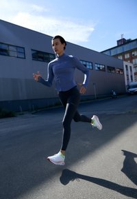 Atleet in een blauwe langemouwen top en zwarte legging die buiten rent, met witte en groene sneakers aan. Stedelijke achtergrond met gebouwen.