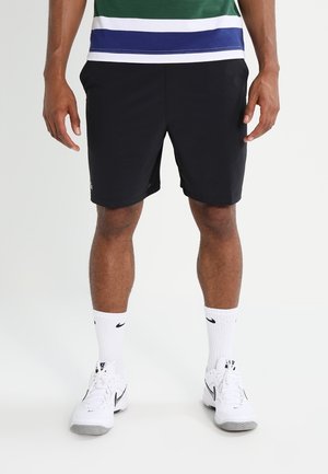 Mann trägt schwarze Sportshorts, weiße Nike-Kurzsocken, weiße Nike-Sneaker und ein grün-weiß-blau gestreiftes Hemd.
