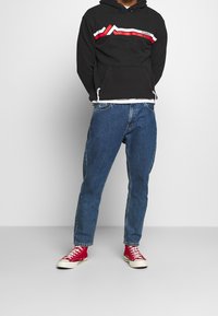 Persona che indossa un felpa nera con strisce rosse e bianche, jeans blu e sneakers rosse alte, in piedi contro uno sfondo semplice.