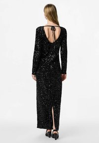 Žena nosi dužu crnu haljinu od šljokica s dugim rukavima, otvorenim leđima vezanim trakama i crnim potpeticama.