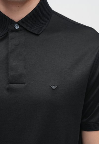 Melna polo krekls no gludas auduma ar teksturētu apkakli, ar 3 pogu plāksnīti un mazu izšūtu logo uz krūtīm.