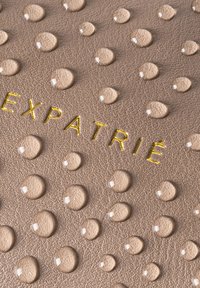 Beige pintas pintatud punktegumas ar augment emblēmunas; "EXPATRIÉ" iegravēts spīdošā zelta burtnīcā.