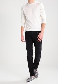 Homme portant une chemise blanche à manches longues, un jean noir texturé et des baskets sans lacets à rayures zébrées, debout devant un fond blanc uni.