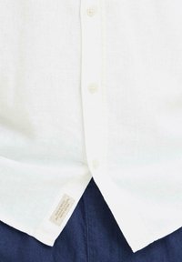 Chemise boutonnée en lin blanc avec un col, comportant une étiquette à l'ourlet, associée à un tissu bleu foncé en bas. Tissu texturé et léger.