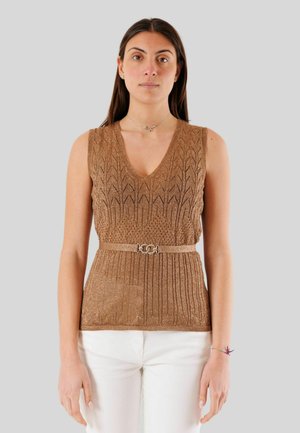 Donna che indossa un top marrone senza maniche in maglia con scollo a V e cintura sottile, abbinato a pantaloni bianchi, in piedi davanti a uno sfondo semplice.