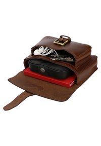 Pochette en cuir marron avec boucle contenant des écouteurs blancs, des clés en métal, un étui noir et un carnet rouge dans deux compartiments.