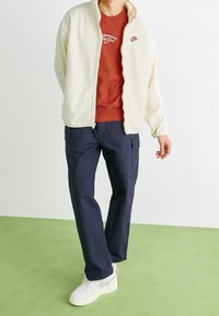 Krämfärgad fleecejacka, rostfärgad t-shirt med vitt logotyp, marinblå cargo-byxor och vita sneakers med texturerade sulor. Modellen står på en grön golv.