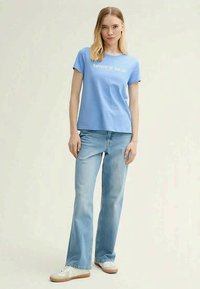 Light blue cotton t-shirt with "HAPPIEST BY THE SEA" printed in white, paired with light blue flared jeans and white sneakers.