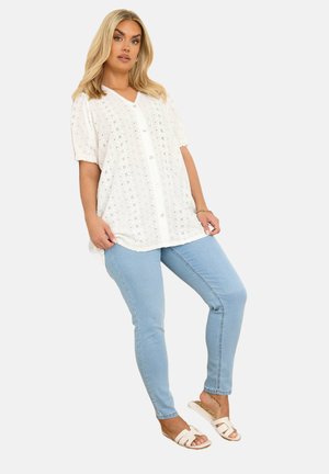 Hvid bluse med knaplukning, øjelet detaljering, korte ærmer og v-hals, parret med lyseblå skinny jeans og hvide slidesandaler.