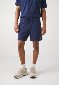 Pantaloni corti blu navy con vita elastica e coulisse, caratterizzati da un piccolo logo bianco. Abbinati a sneaker chiare e calzini bianchi.