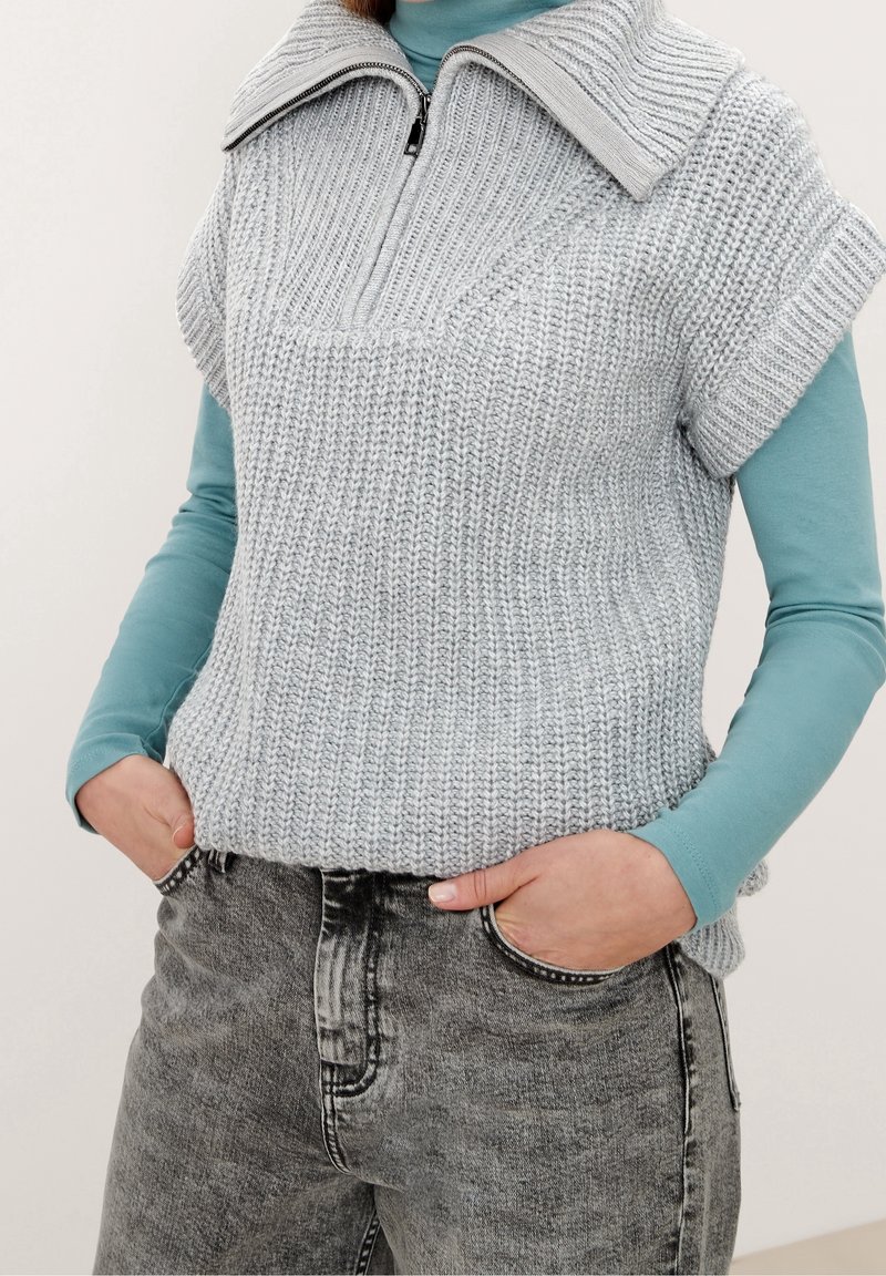 Ljusgråstickad tröja med dragkedja och bred krage, lagd över en teal långärmad skjorta. Bärs med grå slitna jeans.