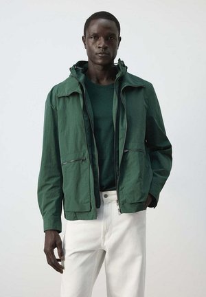 Hombre de piel oscura con chaqueta verde con cremallera y suéter, con pantalones blancos, de pie contra un fondo claro y sencillo.
