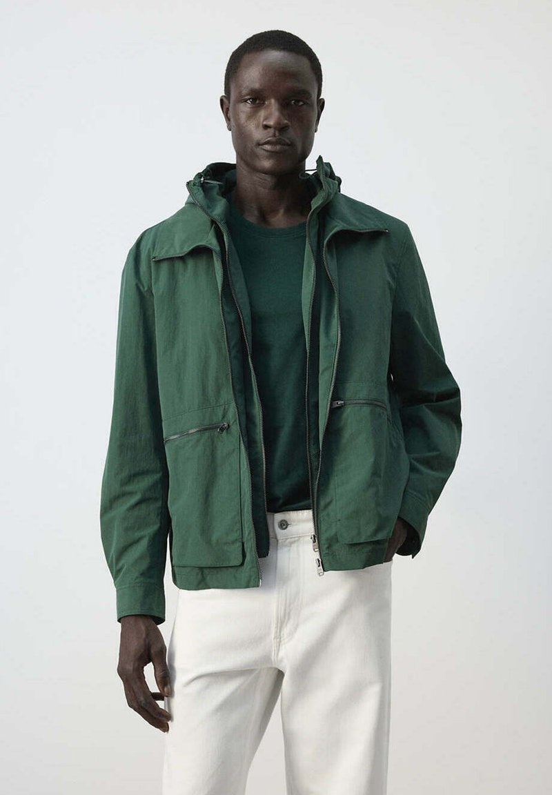 Homme à la peau foncée portant une veste verte zippée et un pull avec un pantalon blanc, debout devant un fond clair et uni.