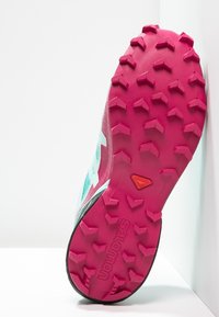 Trailrunning-Schuh mit einer pinken, strukturierten Gummisohle mit aggressiven Stollen; das Obermaterial besteht aus türkisfarbenem Mesh mit schwarzen und weißen Akzenten.