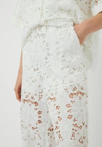 Top bianco in pizzo con motivo floreale e design aperto; presenta maniche larghe e una tasca. Tessuto morbido, leggero e di aspetto trasparente.