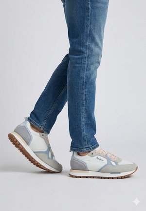 Persona que lleva vaqueros azules y zapatillas blancas-gris-azul con suela de goma, de pie contra un fondo claro y liso.