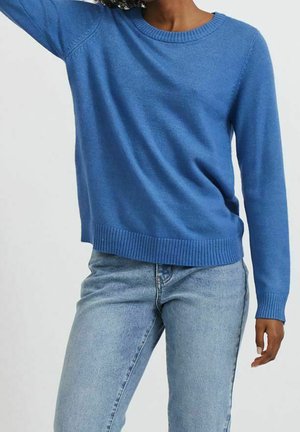 Person iført en blå crewneck-trøje og lyseblå jeans med den ene arm hævet mod en enkel lys baggrund.