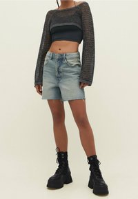 Femme portant un haut court à manches longues en maille noire, un short en denim léger effiloché et des bottes de combat noires à plateforme chunky, se tenant devant un fond uni.