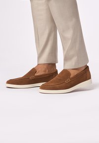 Bruine suede instappers met een witte rubberen zool, voorzien van een gestikte naad aan de voorkant en een minimalistisch ontwerp.