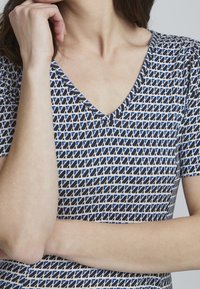 V-ringad topp med ett geometriskt mönster i blått och vitt. Korta ärmar och en mjuk textur förbättrar komfort och avslappnad stil.