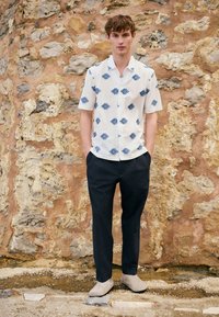 Young man stands with hands in pockets wearing a white short-sleeve shirt with blue patterns, dark pants, and light-colored shoes by a stone wall.