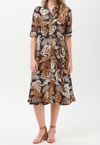 Donna che indossa un vestito midi button-down con stampa tropicale di foglie marroni, beige e nere, con maniche arrotolate e tacchi a blocco beige con cinturino alla caviglia.